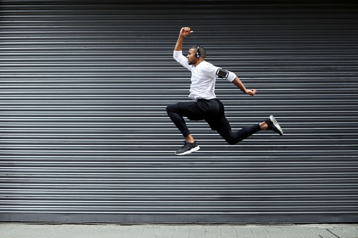 A man leaps into the air, arms and legs bent as if he were running. He's wearing a pair of over ear headphones and a phone strapped to his arm.